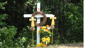 TRAGIC SIGHT: A wooded area on Lake Douglas Road is now adorned with a white cross and some flowers. Handwritten notes were tacked onto the cross, commemorating the three young lives that were lost here in car accident Friday night. Three funeral services were held Tuesday for Adam Goodman, Jordan Harris and Hilary Brock. These services were so crowded they were standing room only. -- Brennan Leathers